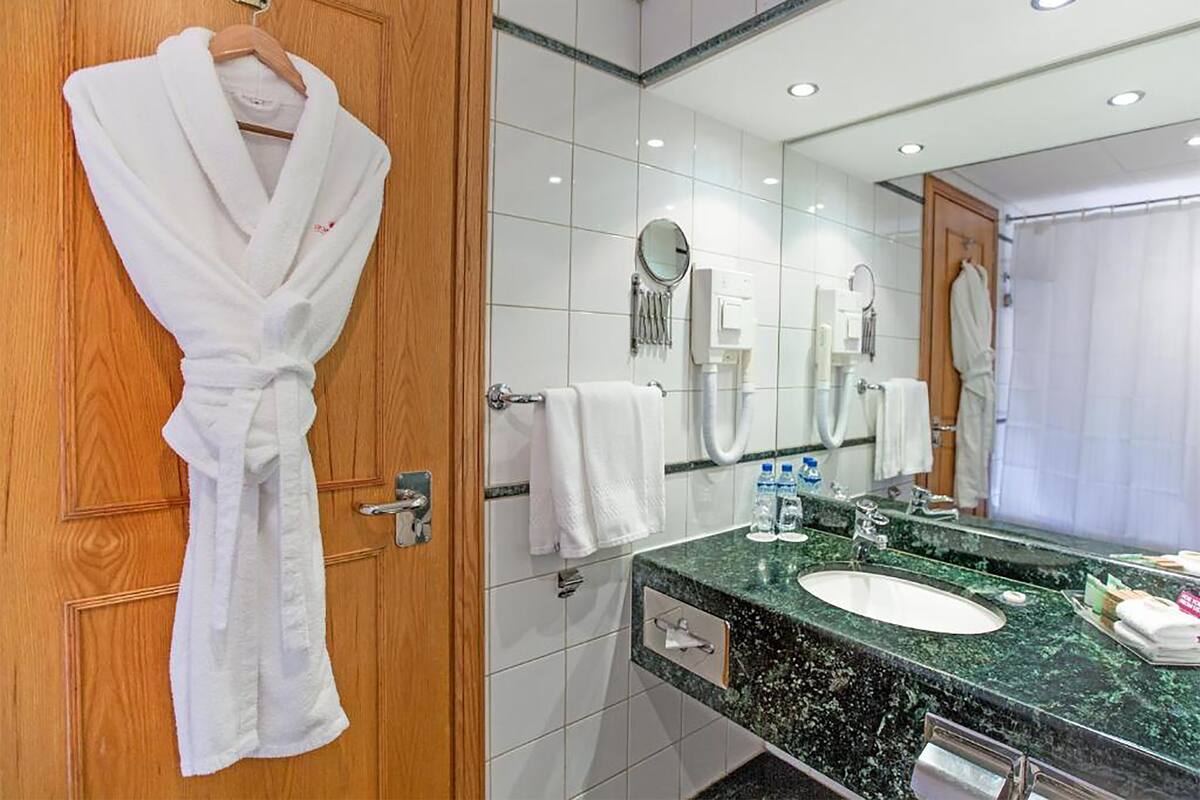 Een hotelbadkamer met een houten deur, een witte badjas hangt aan de deur, een wastafel met spiegel, en toiletartikelen op het aanrecht.