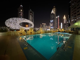Zona de piscina nocturna con una gran estructura iluminada de caligrafía árabe, rodeada de rascacielos urbanos.