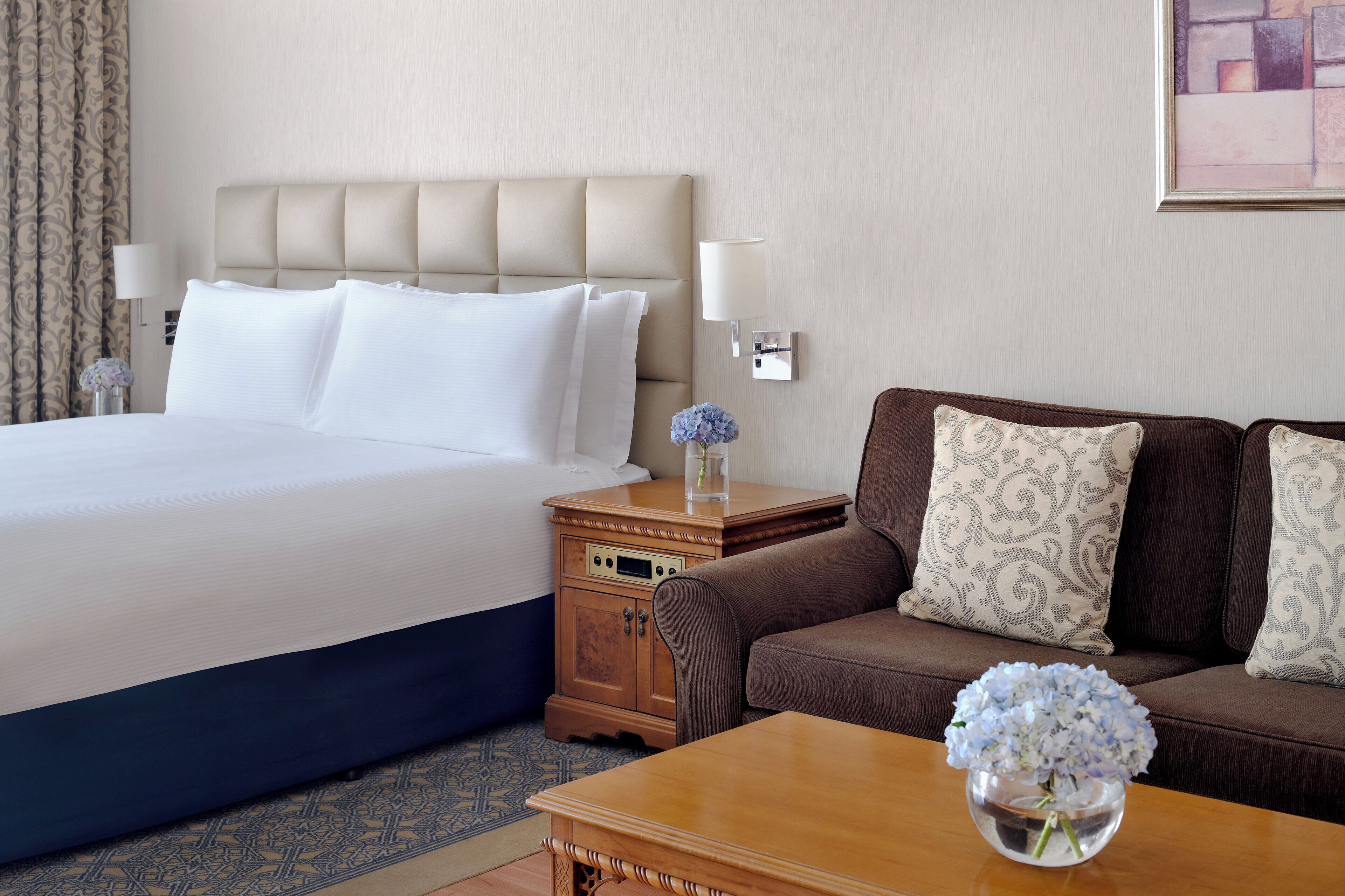 A hotel room with a large bed, a wooden nightstand, a brown sofa, and a small table with a vase of flowers.