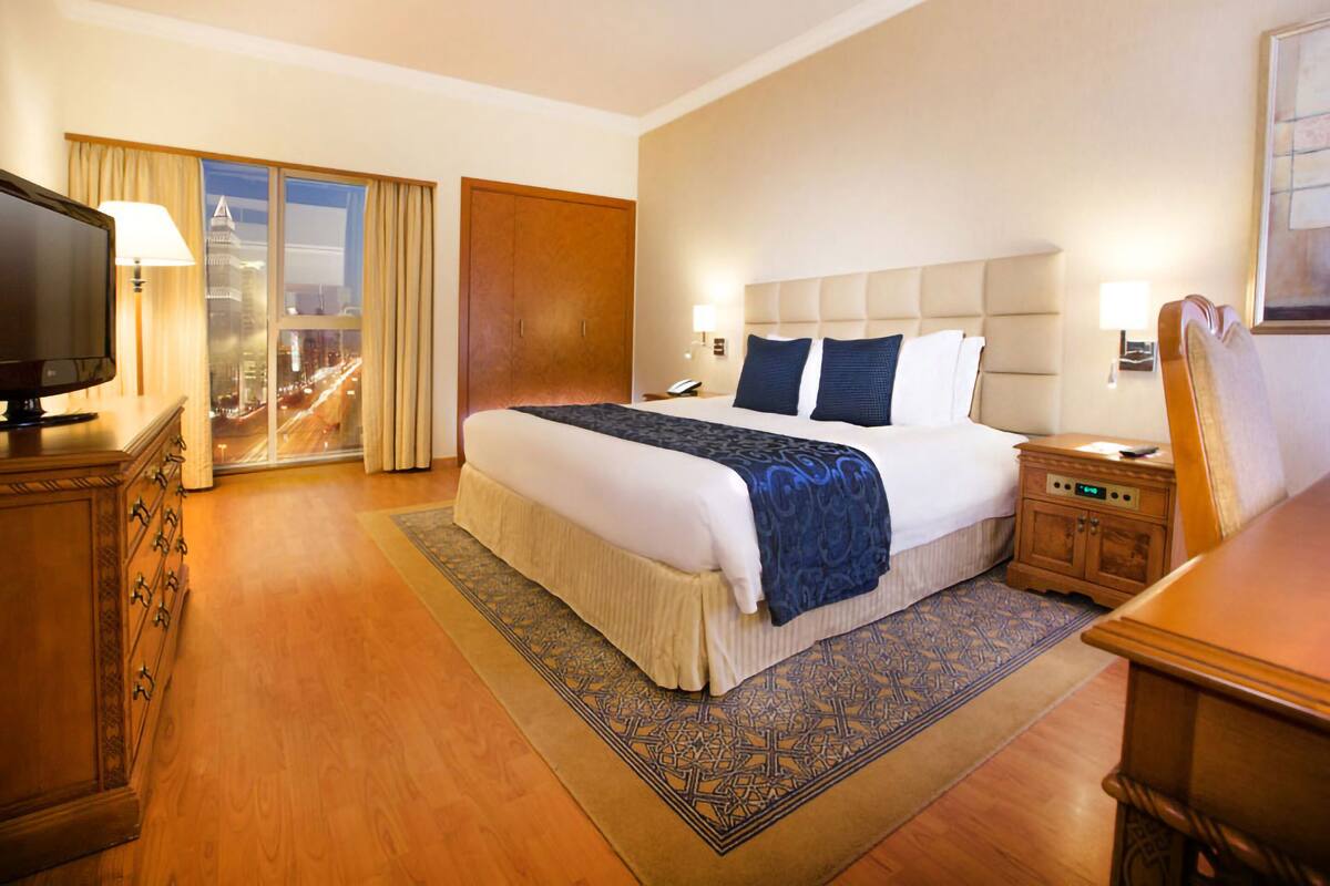 Een hotelkamer met een groot bed, een televisie, een nachtkastje, een houten dressoir en uitzicht op de stad door het raam.