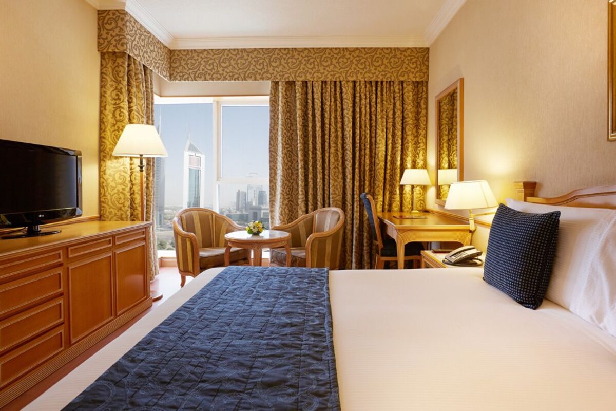 A hotel room with a bed, a TV, a desk with a lamp, a chair, and a view of the cityscape.