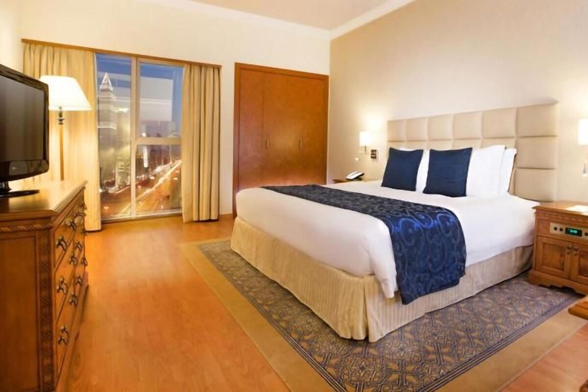 A hotel room with a large bed, a television, a desk, and a view of the city.