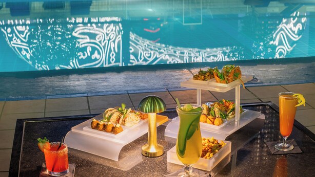 A poolside area with a decorative wall featuring Arabic calligraphy, lounge chairs, and a buffet table with various dishes and drinks.