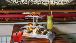 A dining table with food and drinks, a decorative wall with Arabic calligraphy, and a cityscape in the background.