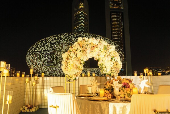 Mesa preparada para cenar con un centro de mesa floral, velas y vistas de edificios altos en la noche.
