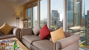 A modern living room with a sofa set, a coffee table with a magazine and a bottle, and a view of a cityscape through large windows.