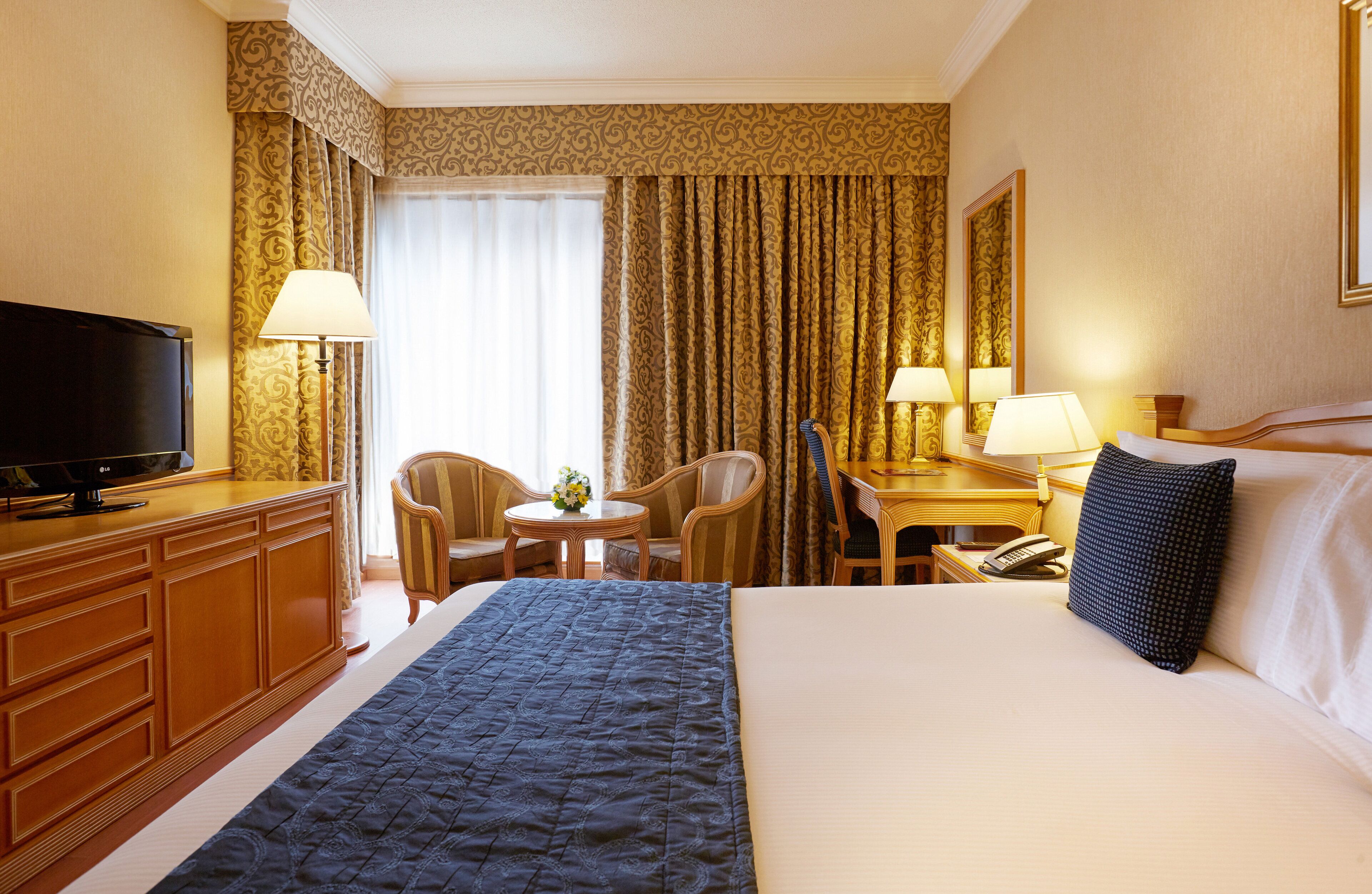 A hotel room with a bed, a TV, a desk with a lamp, a chair, and a small table with a vase.