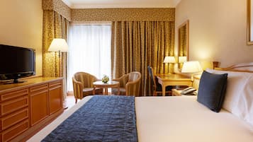 A hotel room with a bed, a TV, a desk with a lamp, a chair, and a small table with a vase.