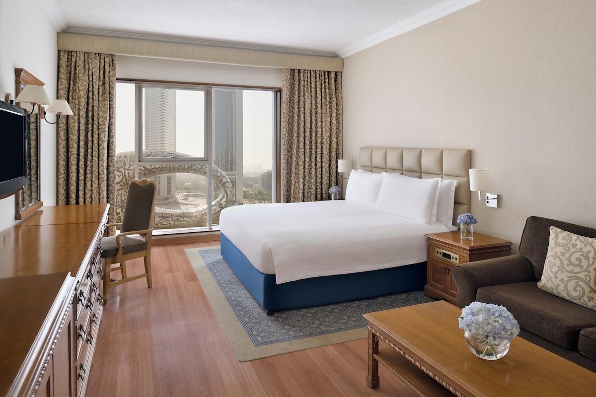 A hotel room with a large bed, a desk with a TV, a chair, a sofa, and a view of a cityscape through the window.