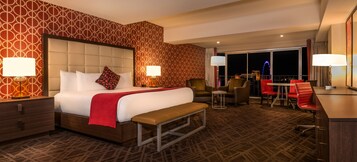 A hotel room with a large bed, two bedside tables with lamps, a bench, a desk with a chair, and a view of the city at night.
