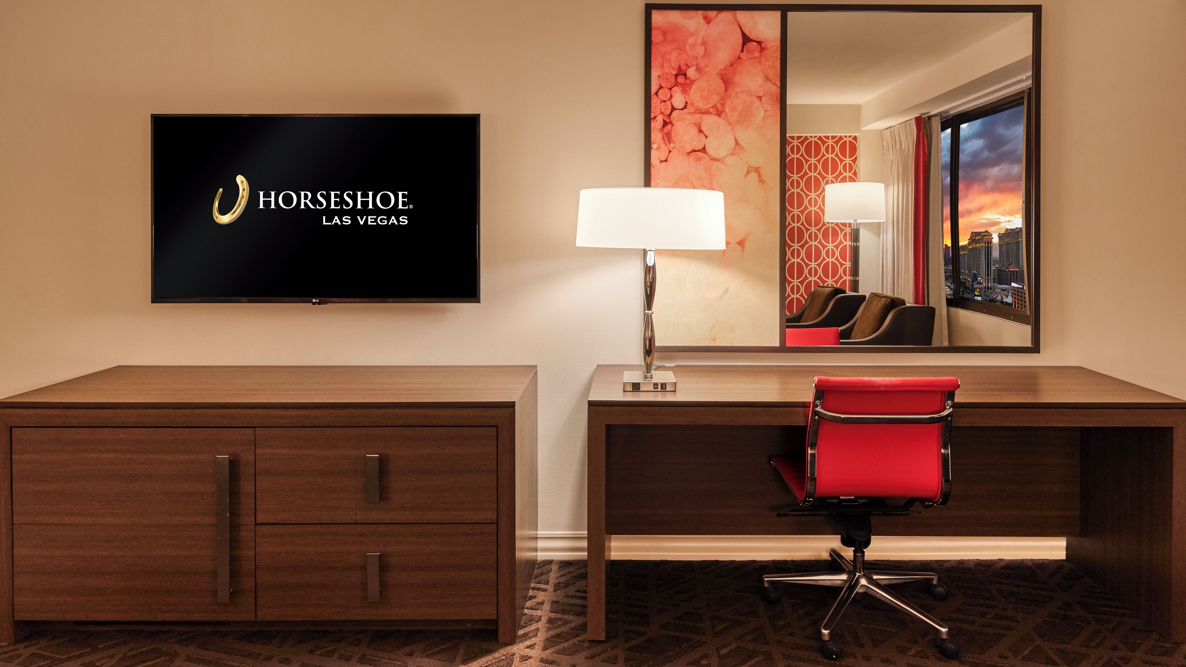 A hotel room with a flat-screen TV displaying the Horseshoe Las Vegas logo, a desk with a lamp, a red chair, and a large mirror reflecting a cityscape at sunset.