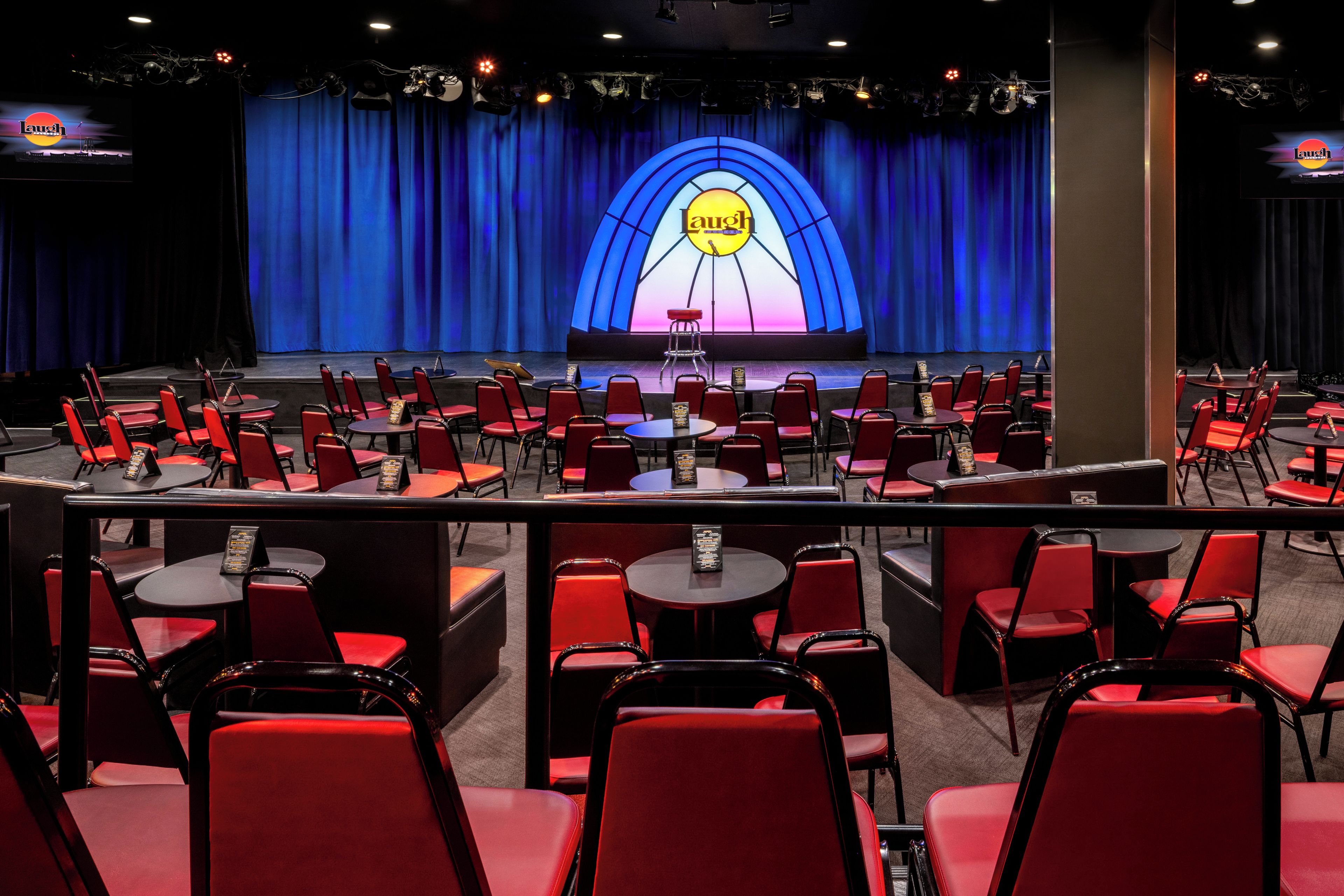 Un salón de conferencias con filas de sillas rojas y mesas, un escenario con un telón de fondo azul y un cartel iluminado que dice 'Laugh'.