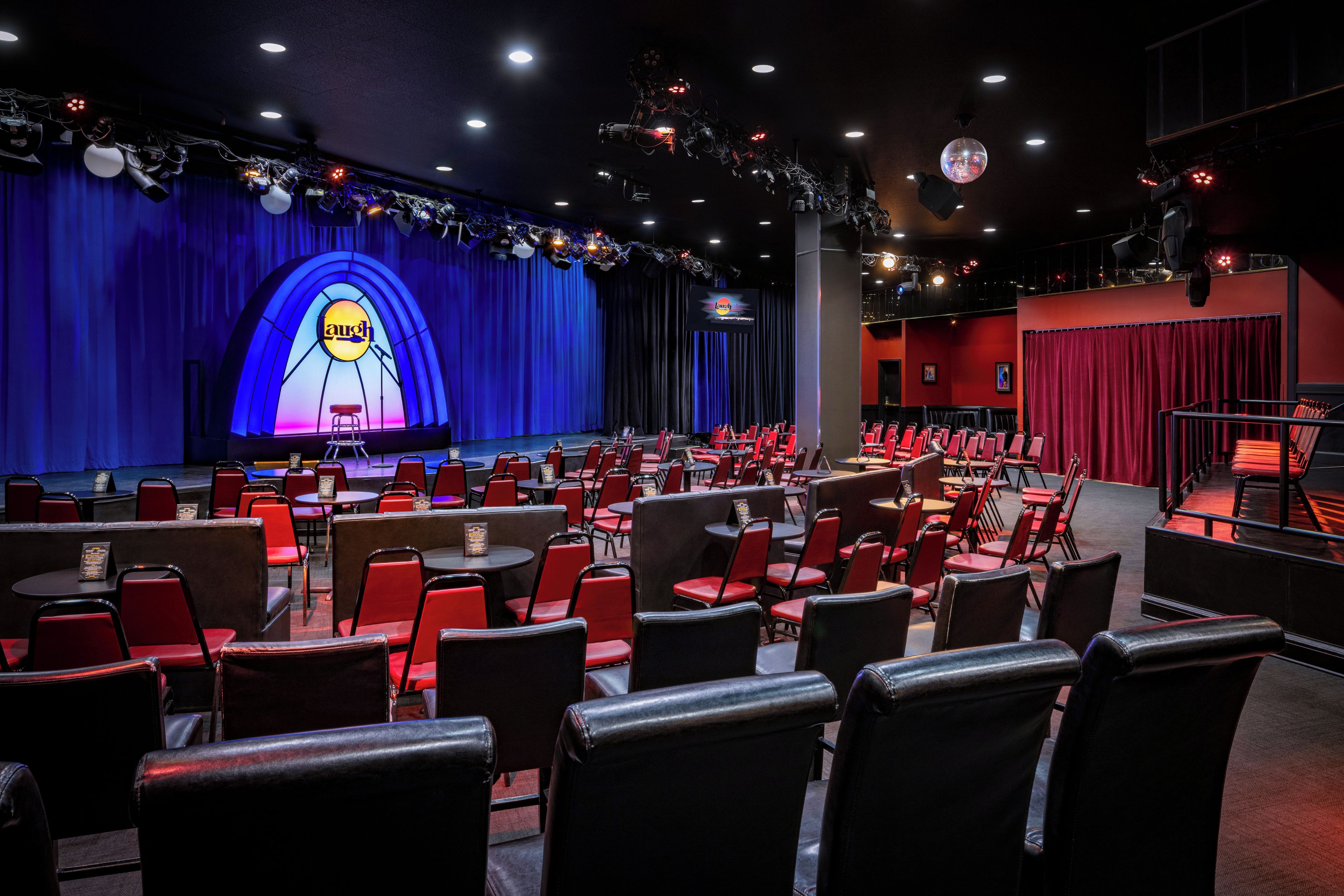 Un teatro con hileras de sillas rojas, un escenario con cortina azul y una pantalla grande que muestra la palabra 'laugh'.