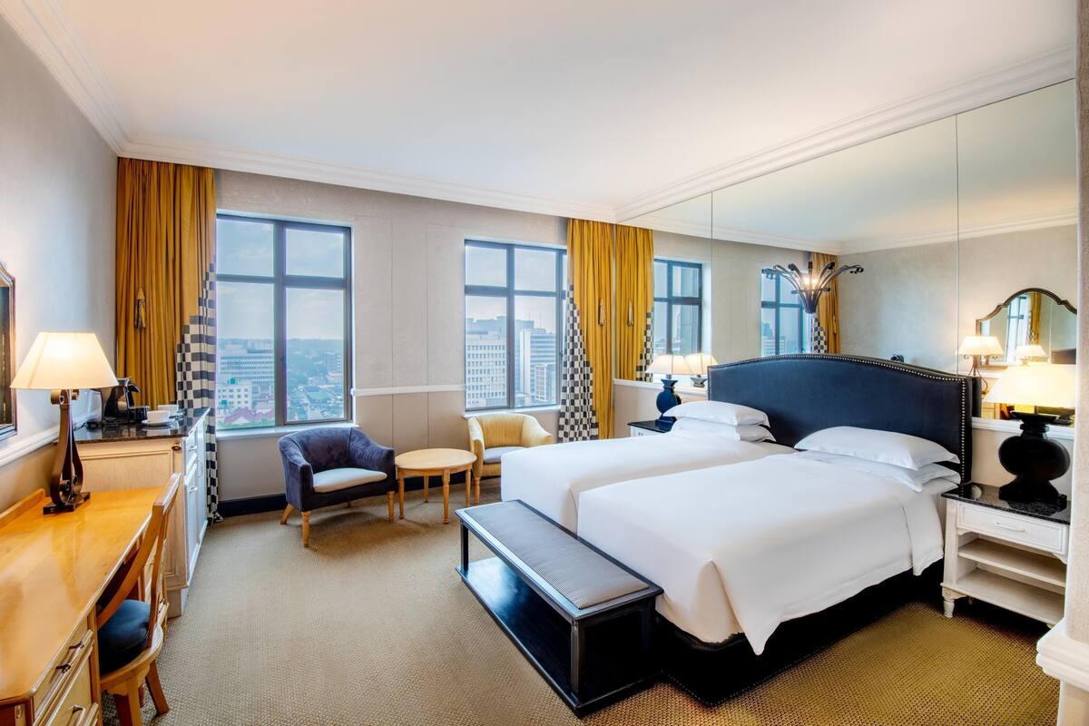 A hotel room with two beds, a desk, a chair, a small table, and a view of the city through large windows.