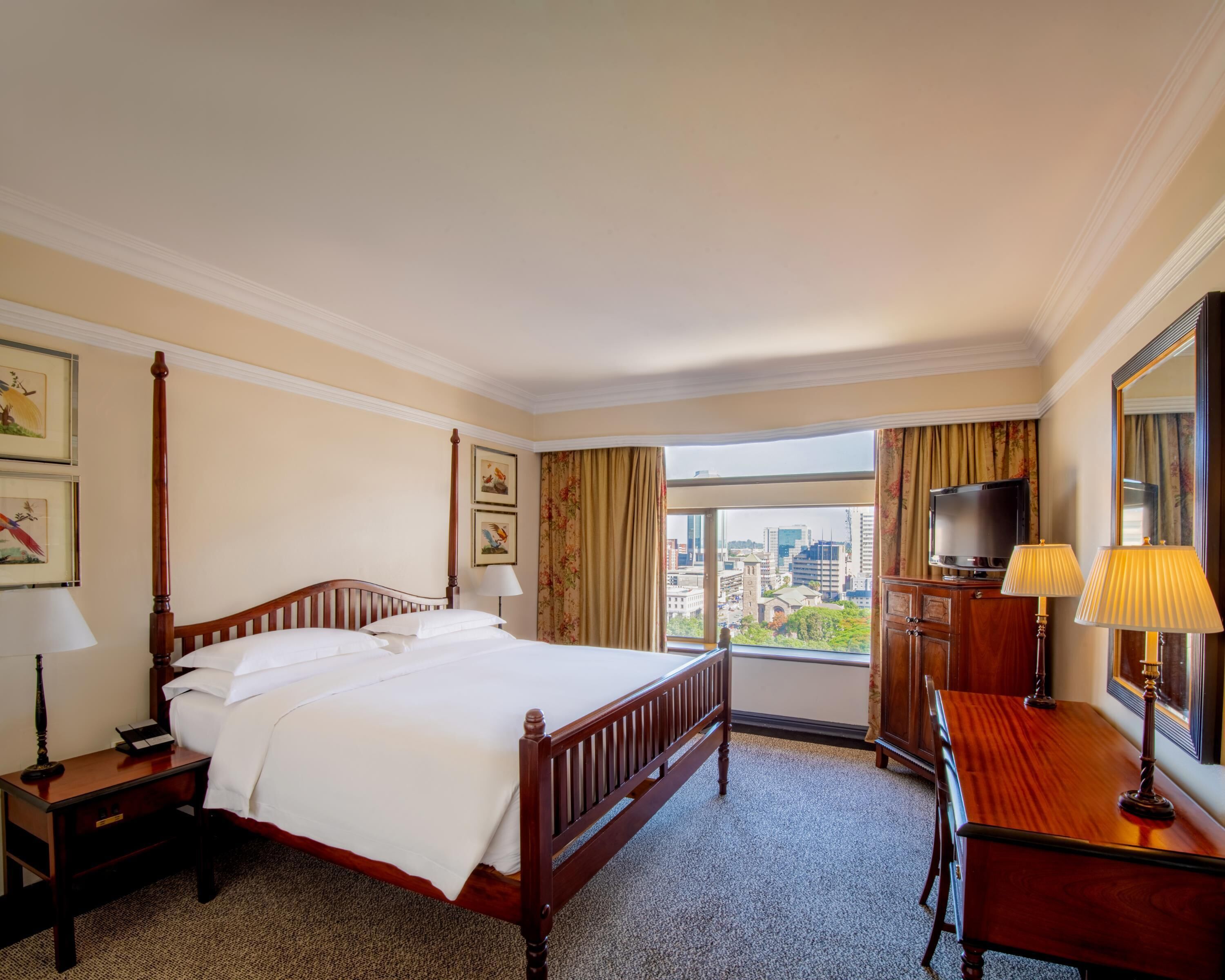 Une chambre d’hôtel avec un lit à baldaquin, un bureau avec une lampe, une télévision et une grande fenêtre donnant sur la ville.