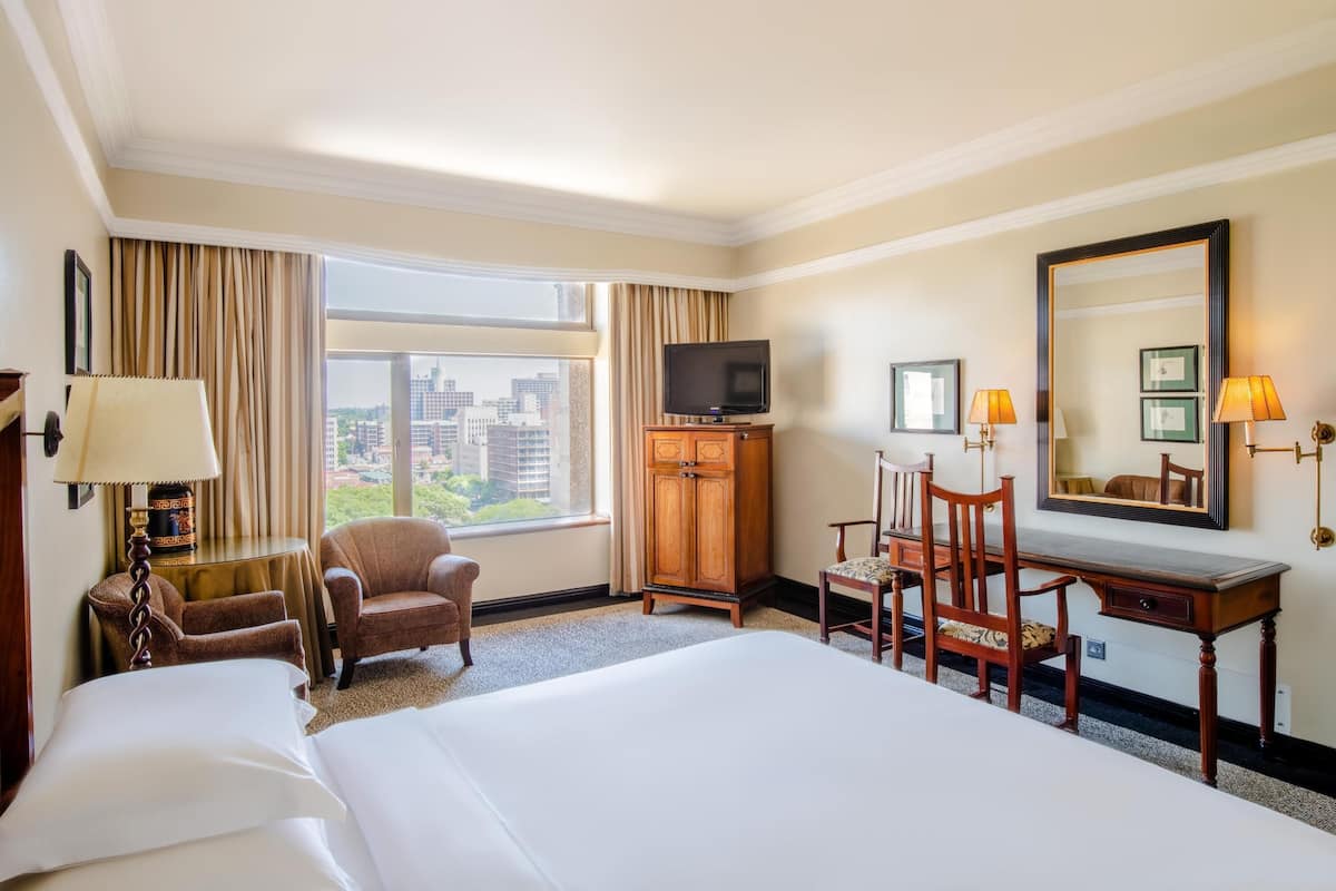 A hotel room with a large bed, two armchairs, a desk with a chair, a television, and a large window offering a city view.