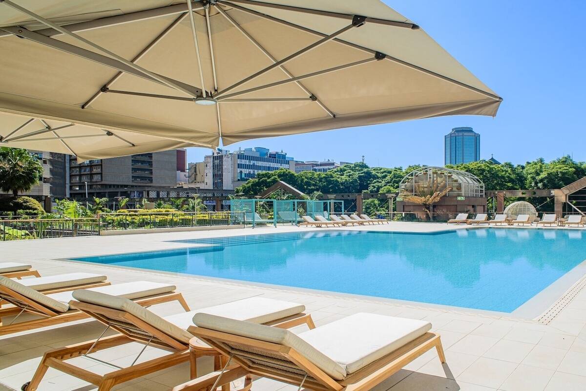 A poolside area with lounge chairs and umbrellas, surrounded by buildings and greenery.