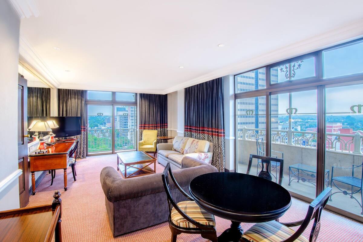 A spacious hotel room with a large bed, a sitting area with a table and chairs, and a balcony with a city view.