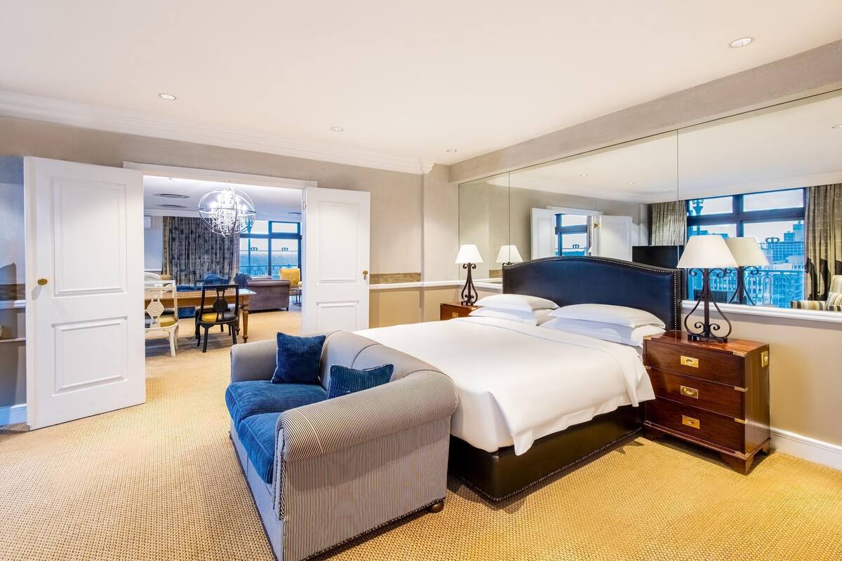 A hotel room with a large bed, a sofa, a dresser, and a mirror. There is a door leading to a dining area.