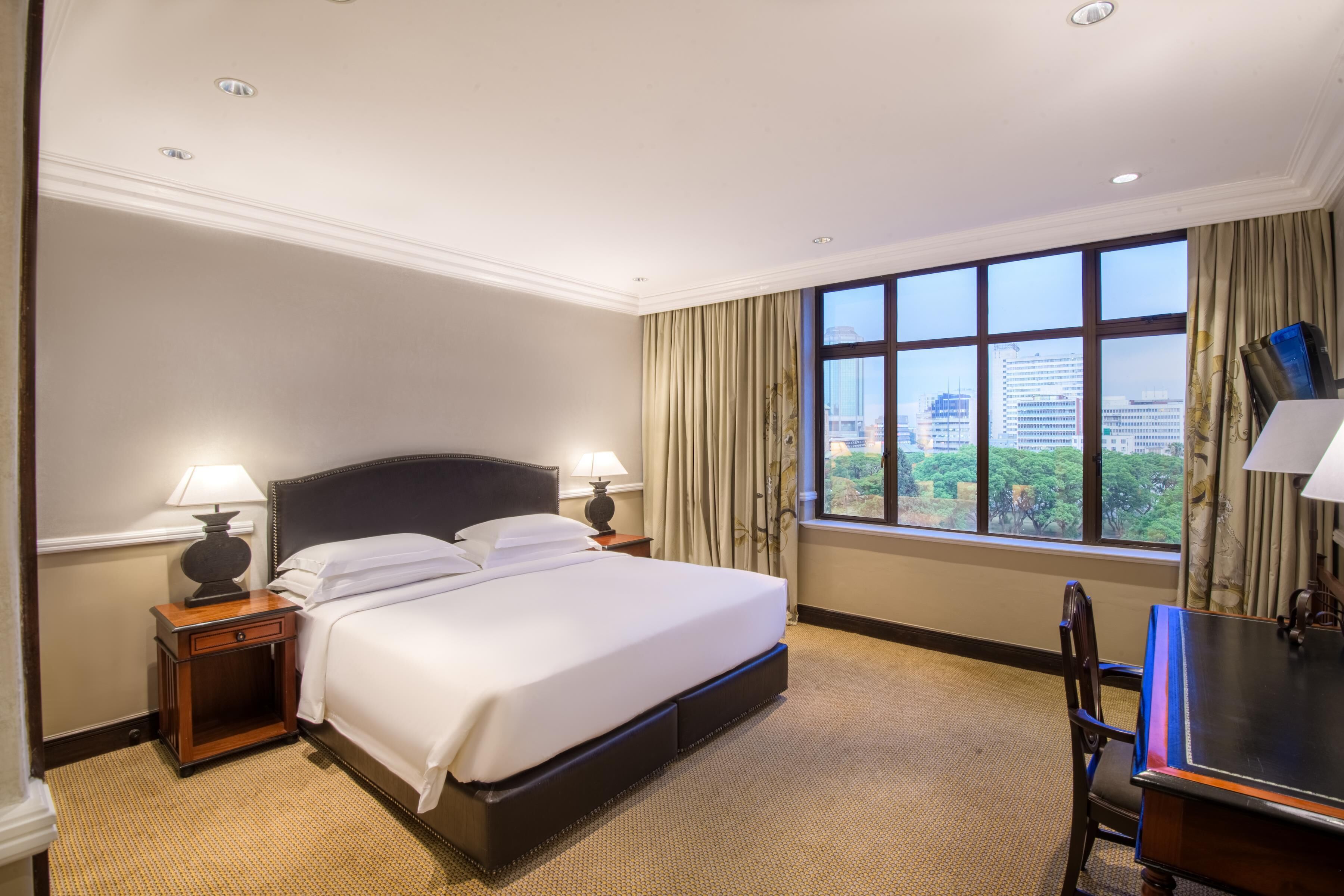 A hotel room with a large bed, two bedside tables with lamps, a desk with a chair, and a large window with a city view.