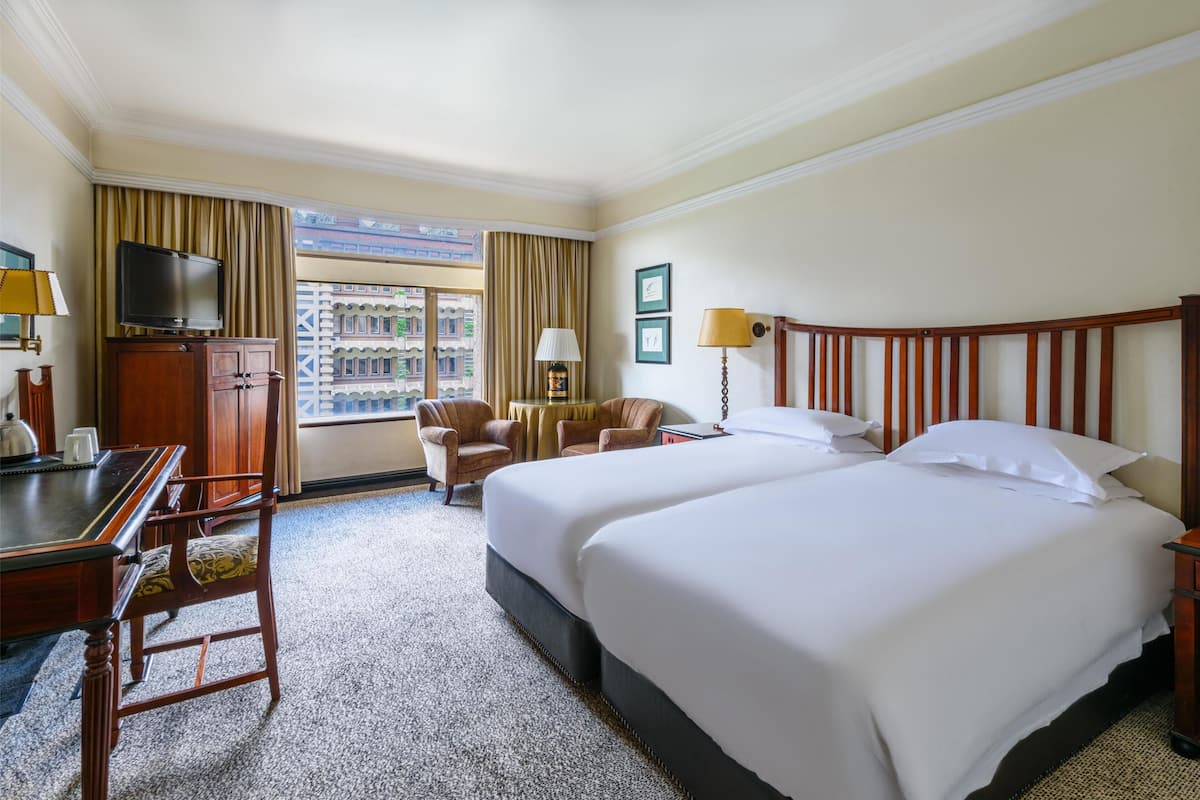A hotel room with two beds, a desk, a TV, and a view of buildings through the window.