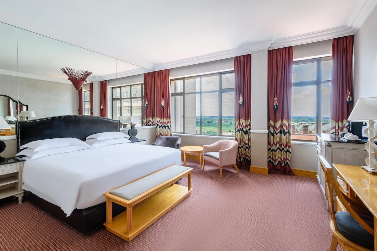 A hotel room with a large bed, a desk, a chair, a small table, and a view of the landscape through the window.