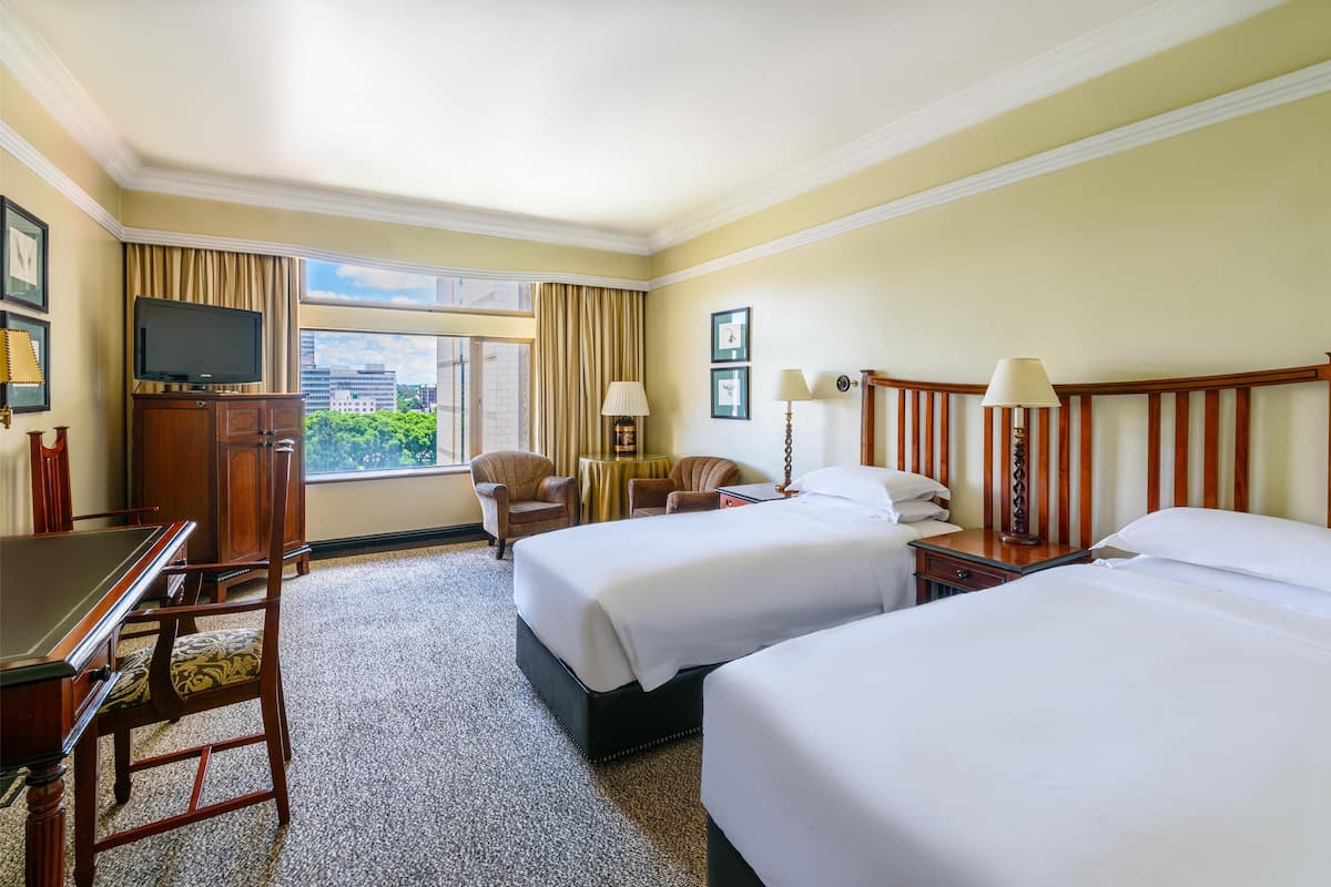 A hotel room with two beds, a desk, a television, and a view of the city.