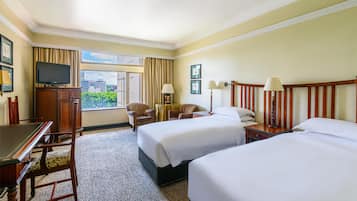 A hotel room with two beds, a desk, a television, and a view of the city.