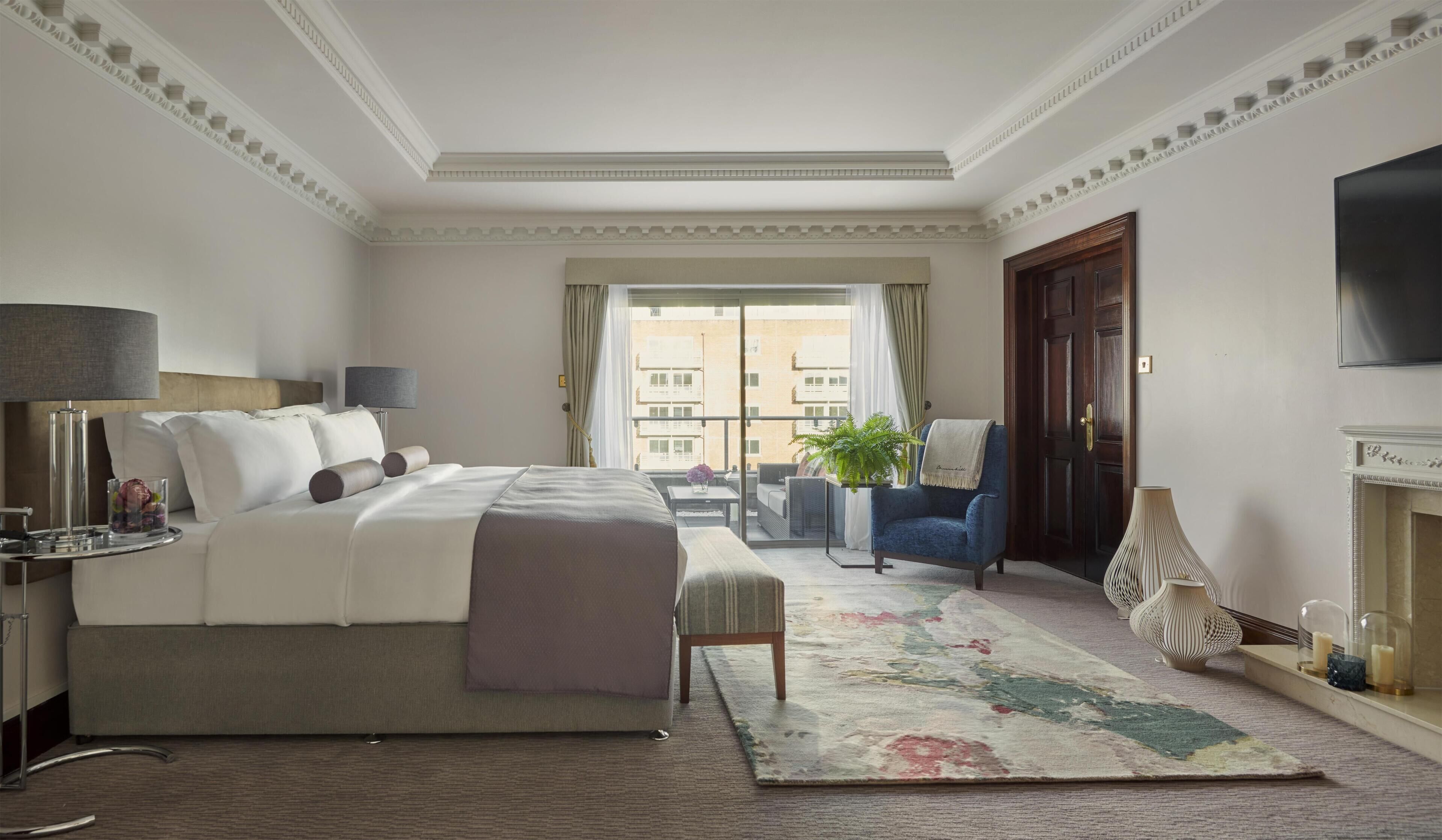A hotel room with a large bed, a sitting area with a chair and a small table, a balcony with a view of buildings, and a fireplace.