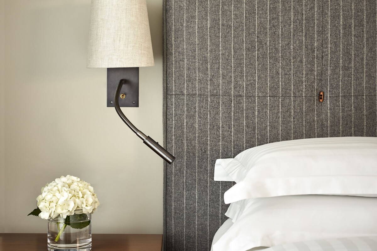 A neatly made bed with white bedding, a wall-mounted lamp with a beige shade, and a small vase with white flowers on a wooden nightstand.