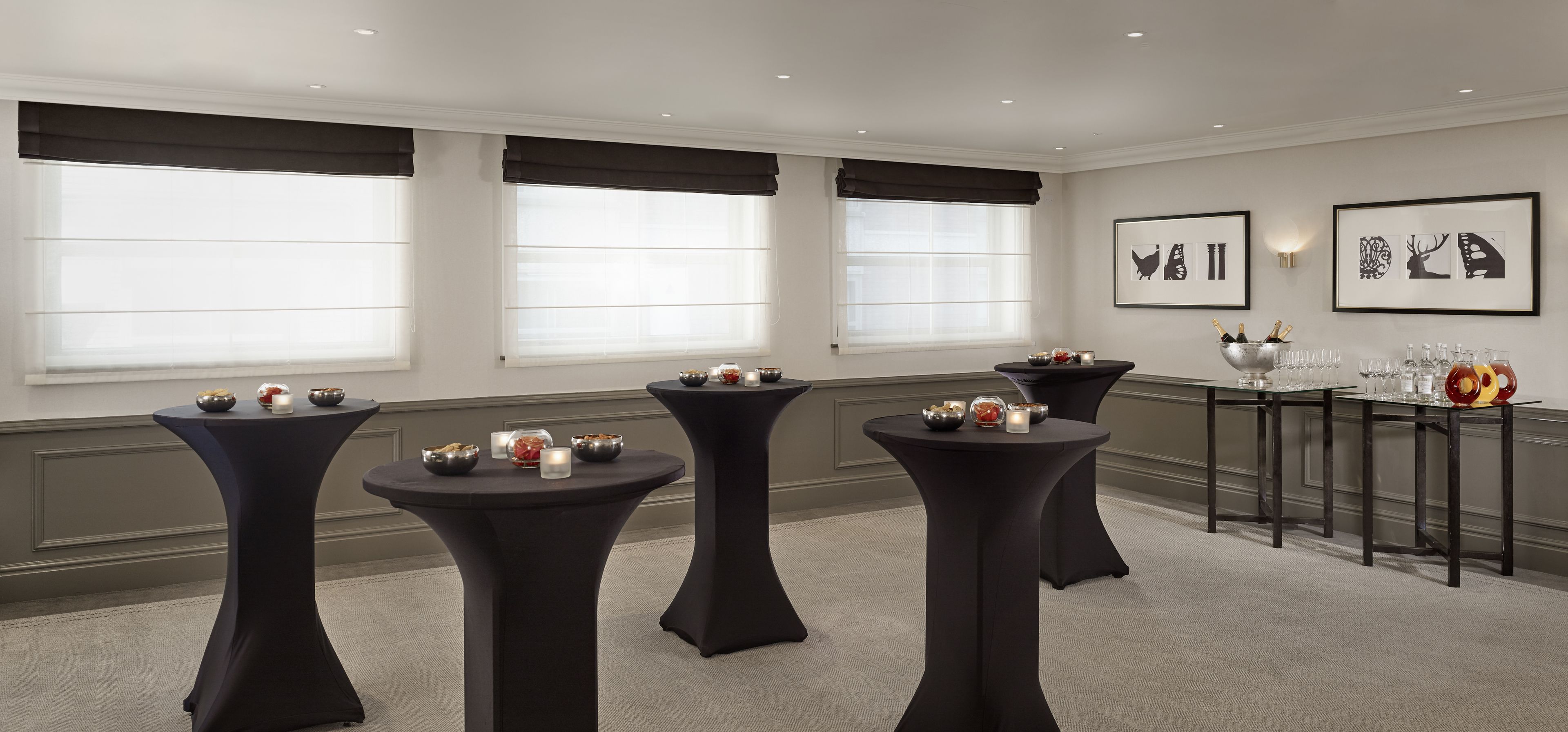 A conference room with four round tables, each with a centerpiece and candles. There are framed artworks on the wall and a buffet table with food and drinks.