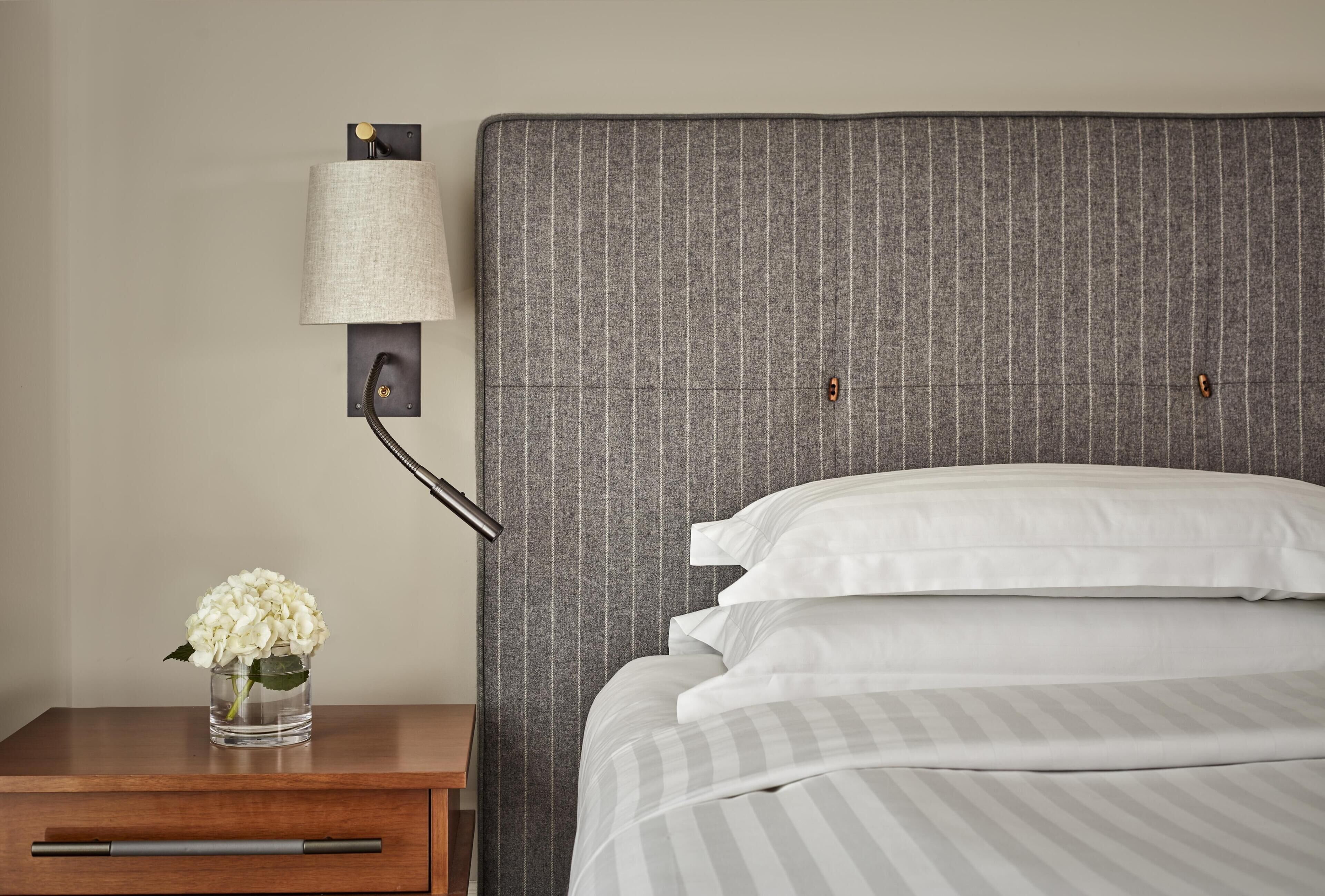 Cama bem arrumada com roupa de cama listrada em branco e cinza, luminária na parede e mesa de cabeceira de madeira com um vaso de flores brancas.