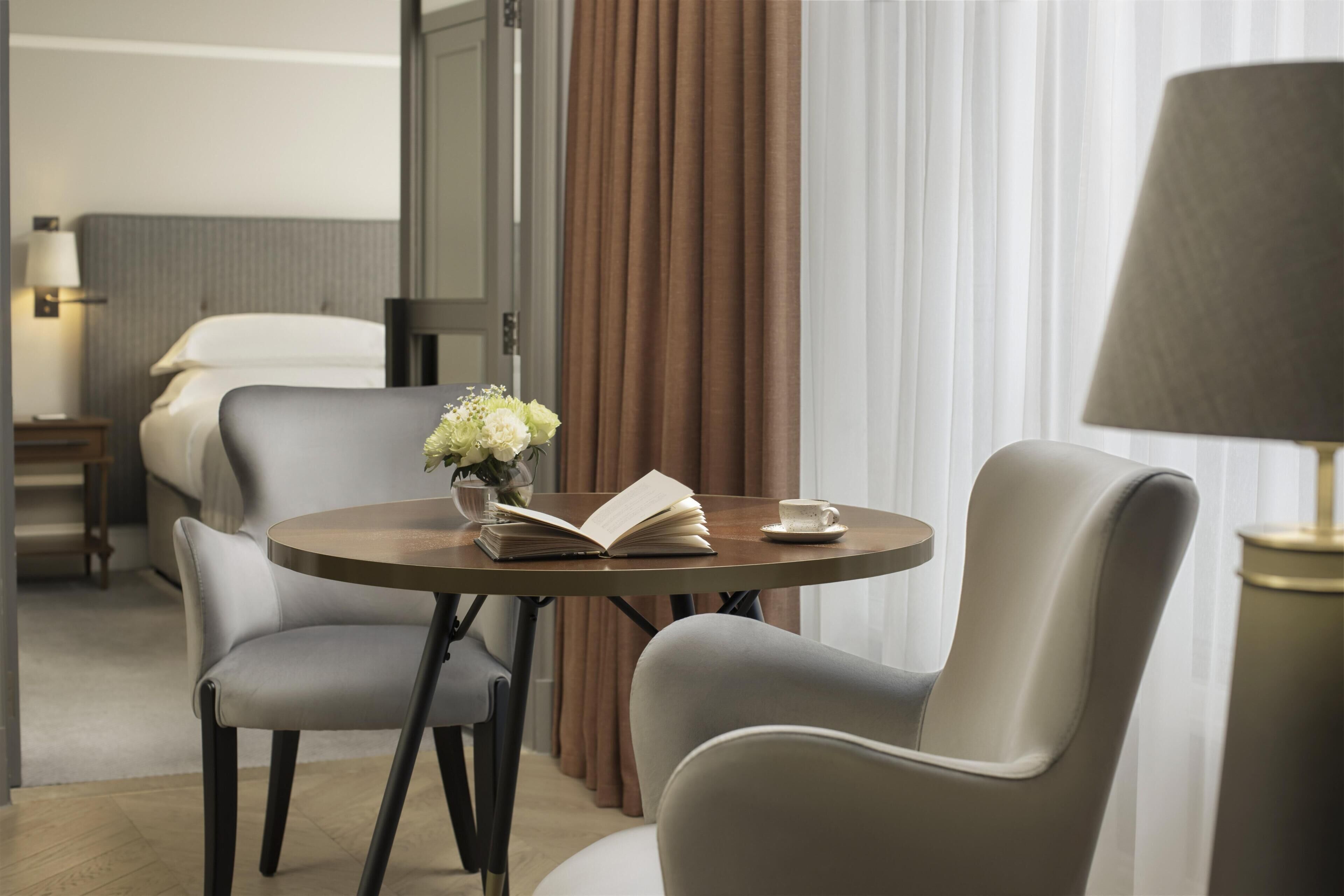 Habitación de hotel con cama, mesita de noche con lámpara, una mesa redonda con un libro y una taza, dos sillas y una ventana con cortinas.