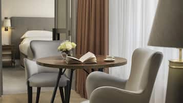 Habitación de hotel con cama, mesita de noche con lámpara, una mesa redonda con un libro y una taza, dos sillas y una ventana con cortinas.