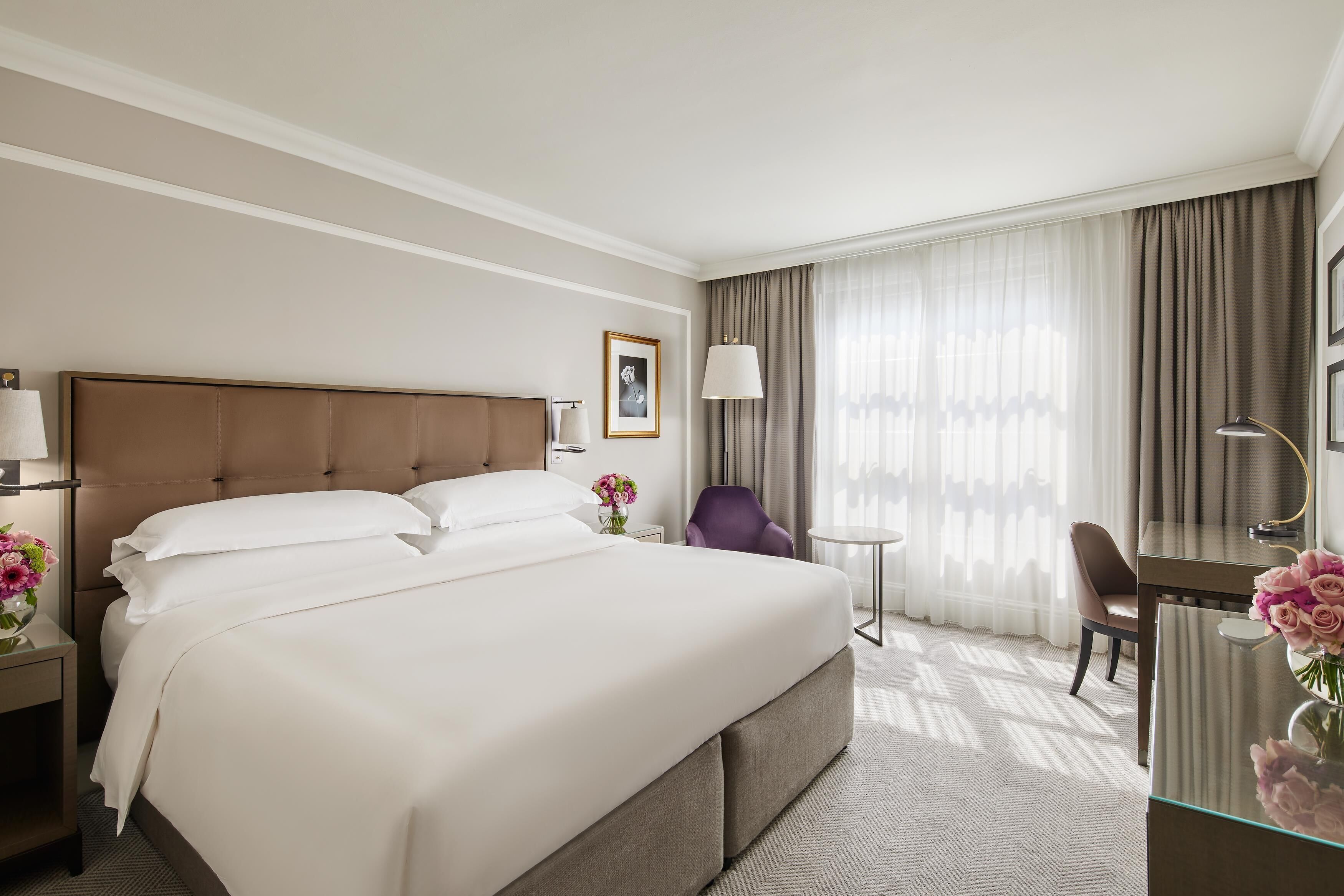 A hotel room with a large bed, a desk with a chair, a small table, and a window with curtains.