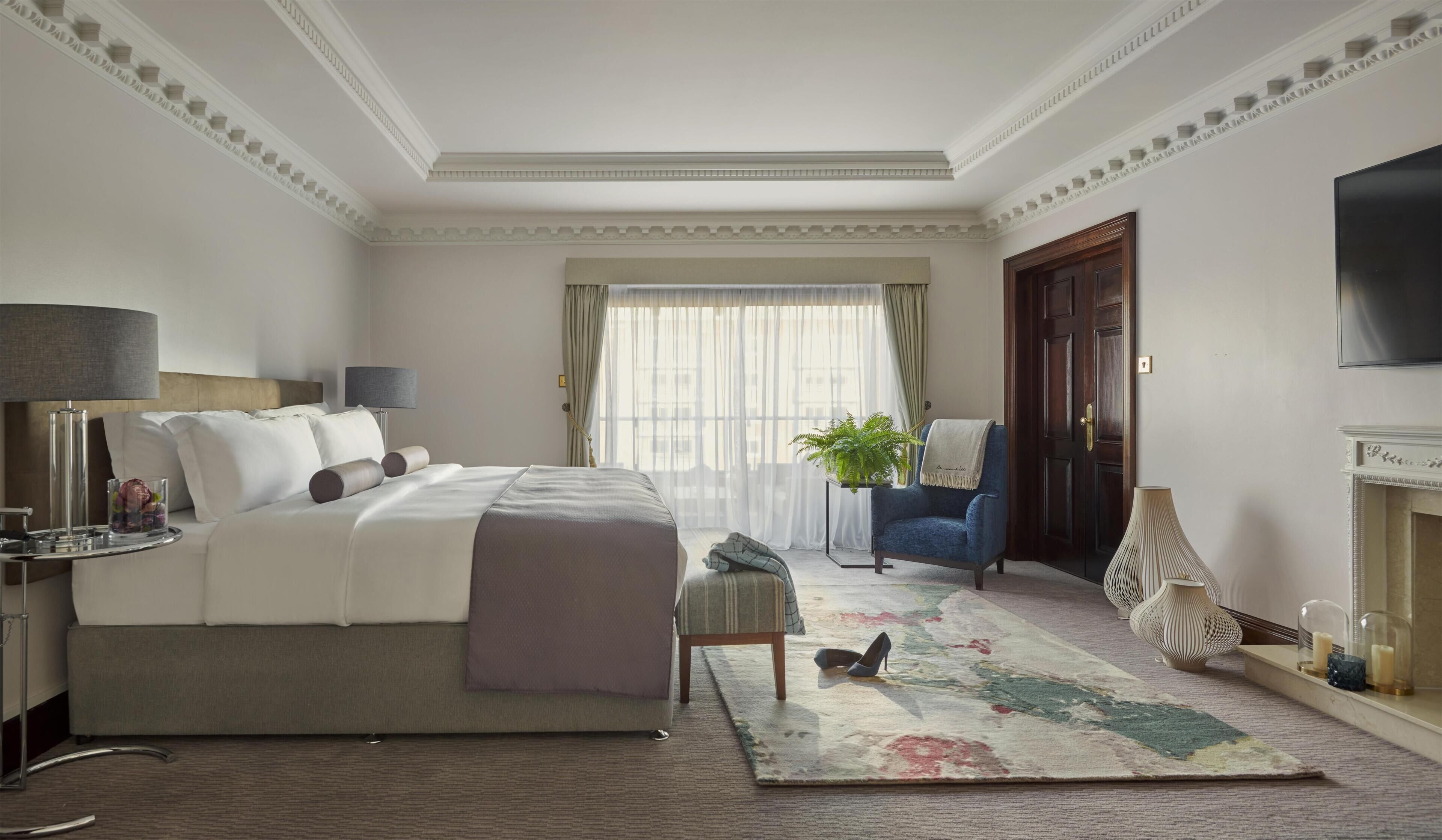 A hotel room with a large bed, bedside lamps, a small table with a lamp, a chair, a potted plant, a rug, and a television mounted on the wall.