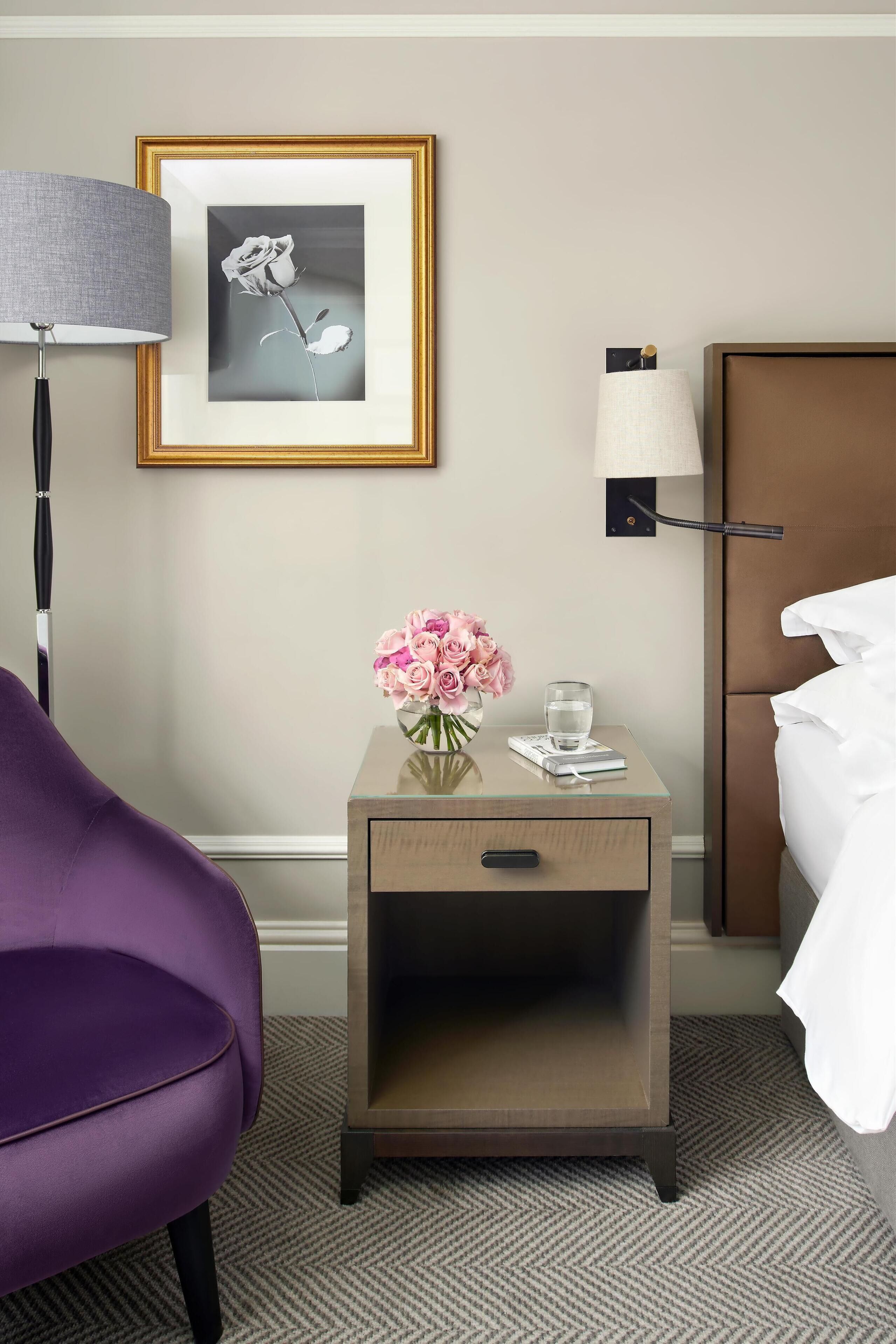A hotel room with a bed, a nightstand with a lamp, a purple chair, a framed picture on the wall, and a vase with pink flowers.