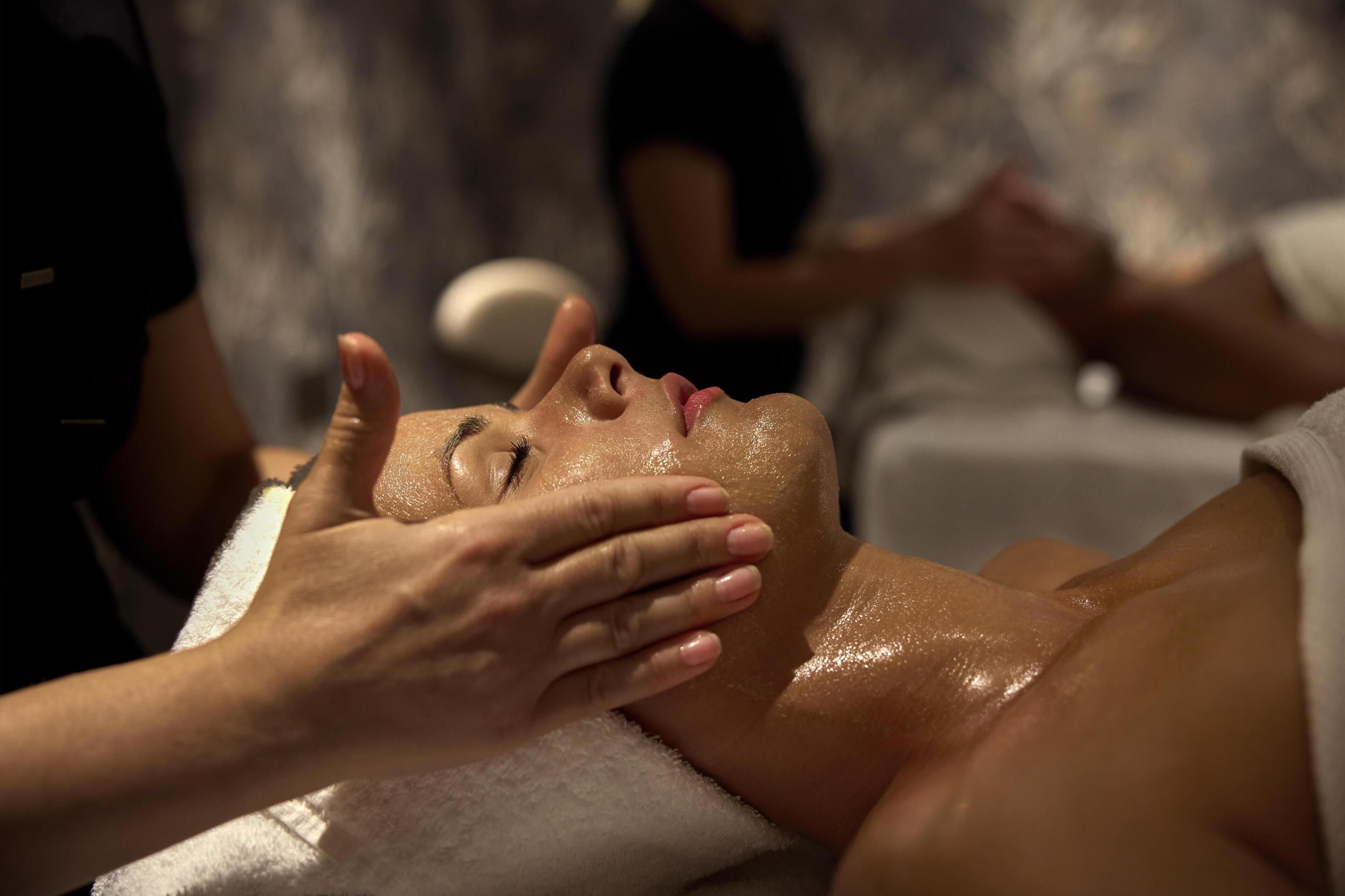 A person receiving a massage with hands on their face, another person in the background, and a white towel.