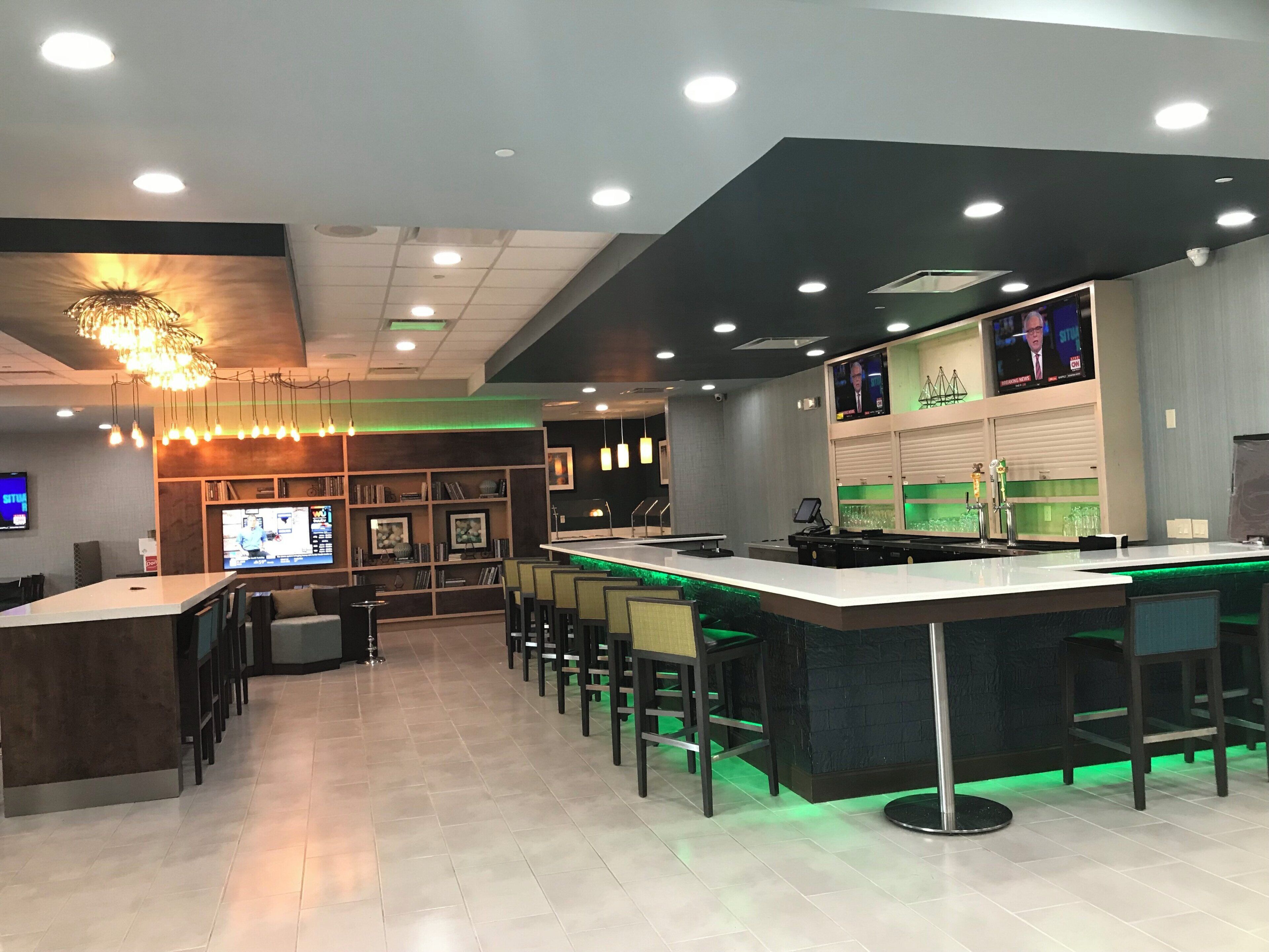 Zona bar moderna con un lungo bancone, diverse opzioni di seduta e uno schermo televisivo.