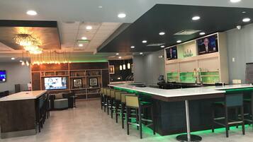 Zona bar moderna con un lungo bancone, diverse opzioni di seduta e uno schermo televisivo.