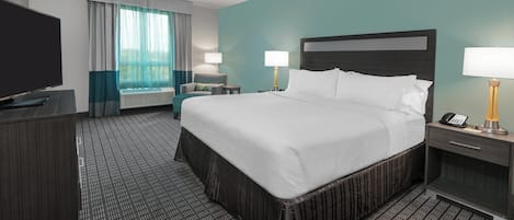 A hotel room with a large bed, a desk with a flat-screen TV, a chair, two bedside lamps, and a window with curtains.