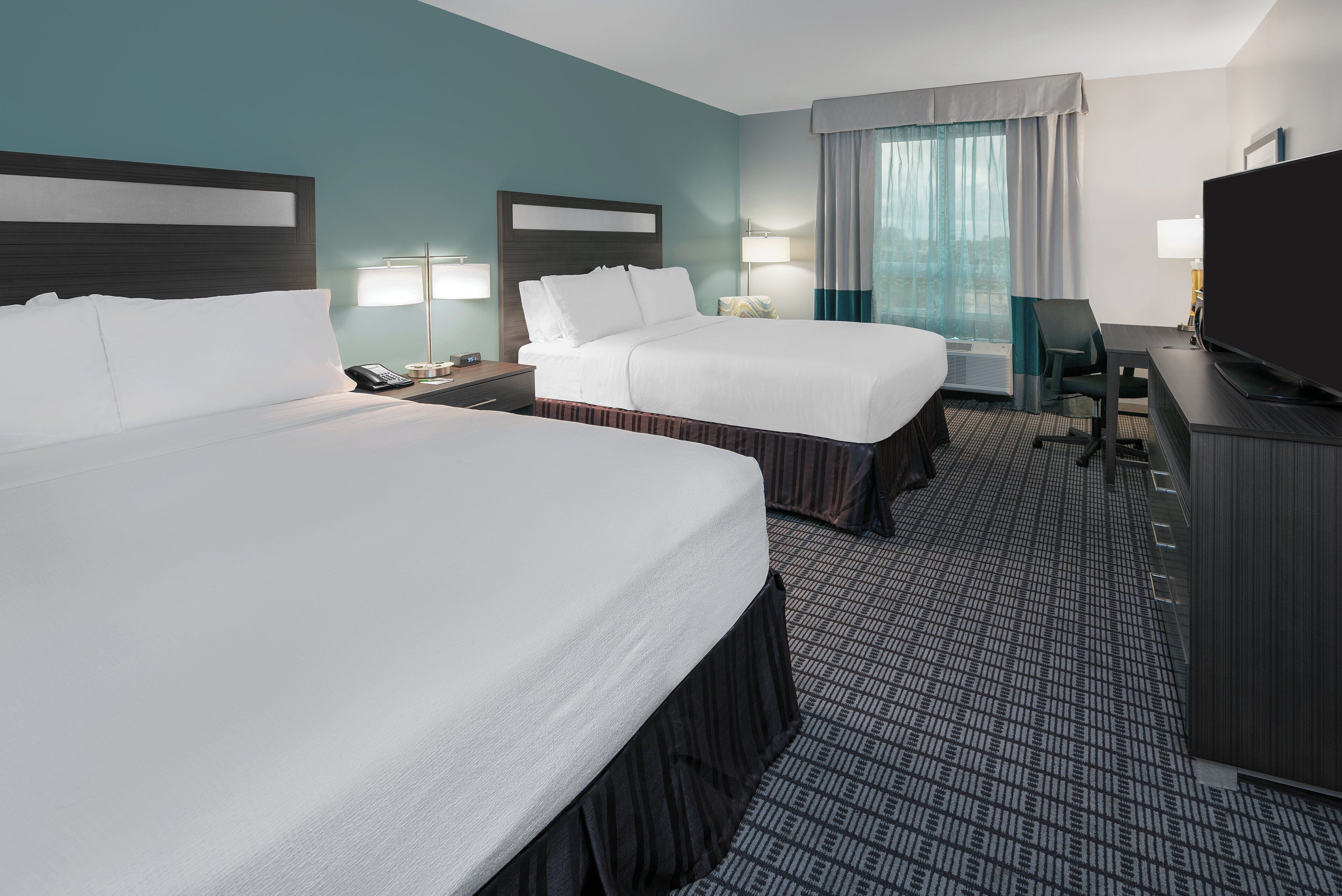 A hotel room with two beds, a desk, a TV, and a window with curtains.