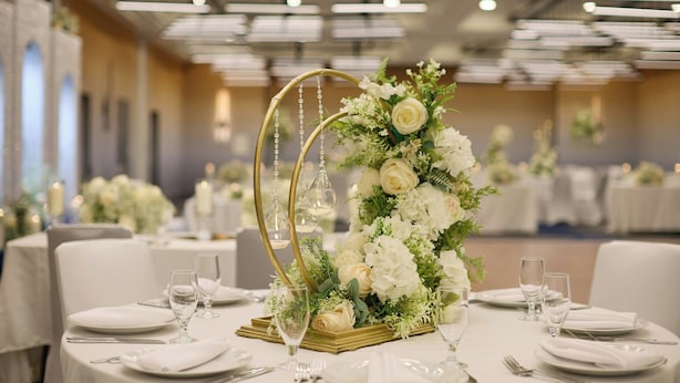 Un salón de banquetes con mesas redondas dispuestas para un evento formal, con centros de mesa florales blancos y detalles dorados.