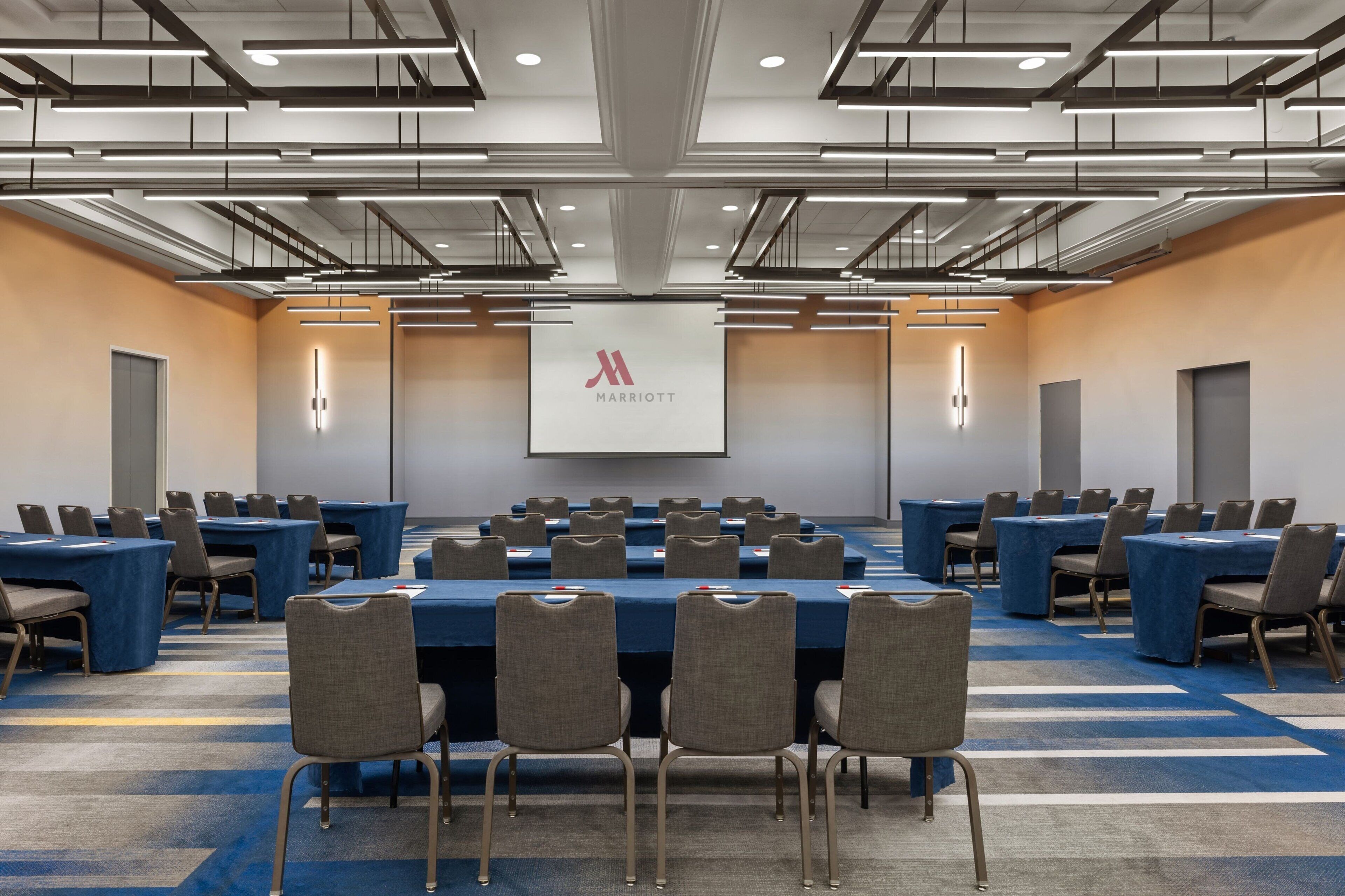 A conference room with multiple tables and chairs, a large screen, and a Marriott logo on the wall.
