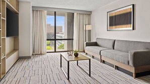 Una habitación de hotel moderna con sofá, televisor de pantalla plana, mesa de centro y un amplio ventanal con cortinas.