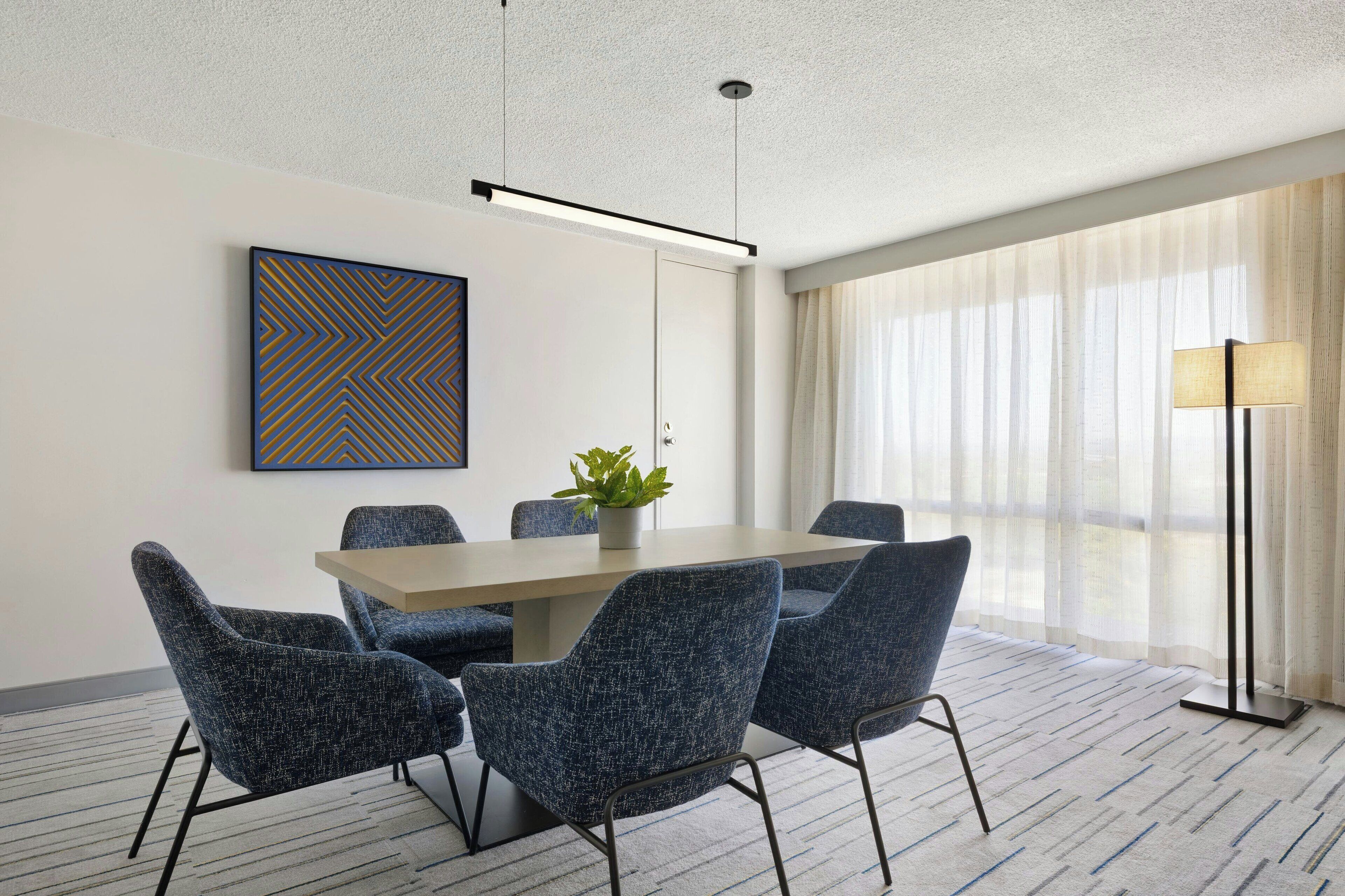 A conference room with a rectangular table, four upholstered chairs, a potted plant, a framed artwork, and a floor lamp.