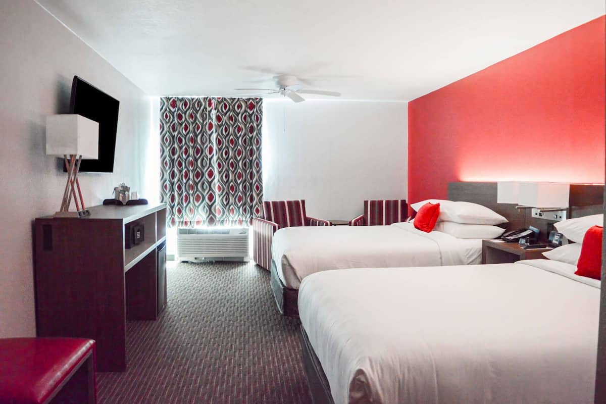 Ein Hotelzimmer mit zwei Betten, einem Schreibtisch, einem Fernseher und einer roten Wand als Gestaltungselement.