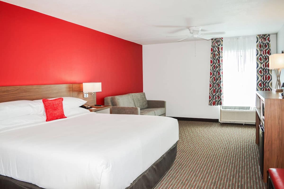 A hotel room with a large bed, a desk with a lamp, a chair, and an air conditioning unit.