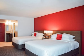 A hotel room with two beds, a red accent wall, a mirror, a sink, and a nightstand with a lamp.