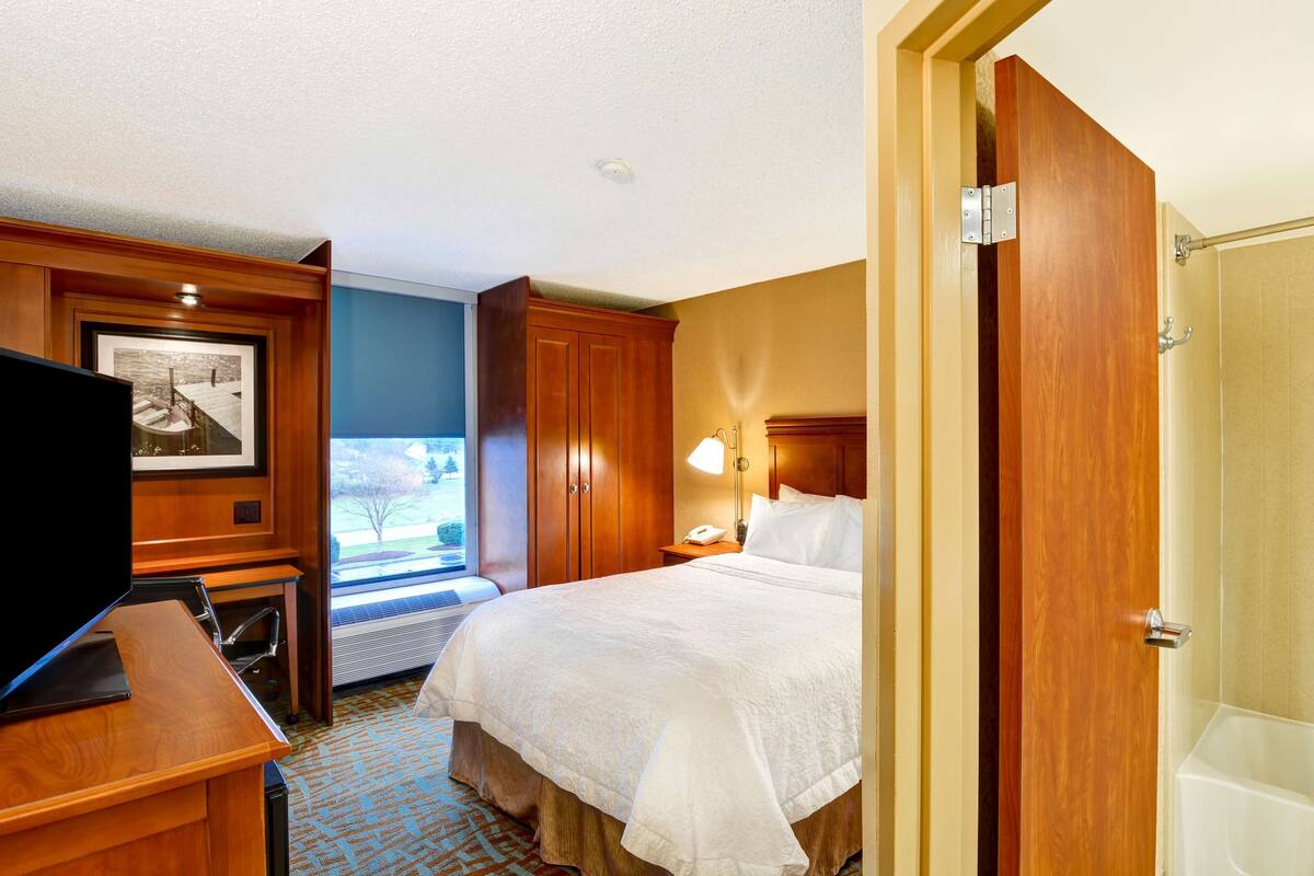 Ein Hotelzimmer mit Bett, Schreibtisch mit Fernseher, Fenster, Tür und einem Badezimmer mit Badewanne.